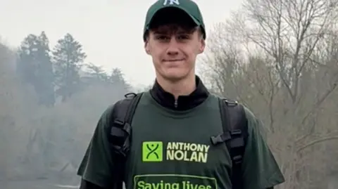 A young man standing on a bridge over a river. He is wearing a green New York Yankees baseball cap and a green t-shirt which reads: "ANTHONY NOLAN - Saving lives through stem cells." He has a rucksack on his back and is holding what looks like a map.