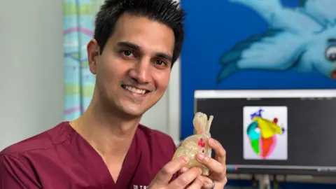 Picture of the doctor holding a replica 3D heart. The heart is made of transparent plastic and fits in his hands. The doctor has short black hair, spiked up, and wears a burgundy scrubs top.