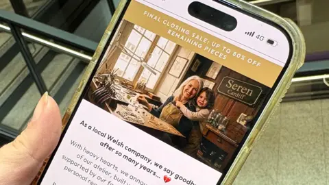 A mobile phone being held in someone's hand. On the phone is an image of an older woman with an apron sitting at a workshop desk, with a young girl hugging her. A sign saying 'Seren Cardiff' can be seen in the background. 