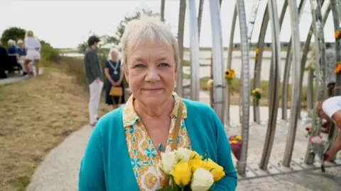 Tim Everest/BBC Caroline Schilt pictured in front of the Old Shoreham Toll Bridge in Shoreham. She has short white hair and is wearing a blue cardigan and a blue and yellow patterned blouse. She is holding a bouquet of flowers.