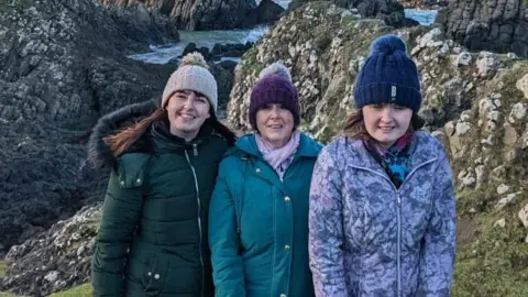 Eileen Mitchell Two women and a teenager pose on top of rocks by the sea. They are all smiling and wearing bobble hats, gloves and coats. The sea is calm behind them.