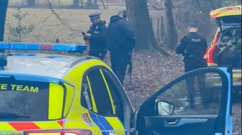 Three police officers in black uniforms, carrying black rifles, stand on a track next to a field. 