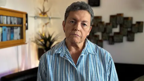 Gemma Dillon/BBC A woman stood up in a room, staring straight ahead. She has short, grey hair and is wearing a blue and white striped shirt. In the background are items hanging on a wall.