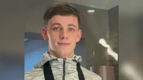 A teenage man with short brown hair and wearing a grey zipped jacket is smiling.