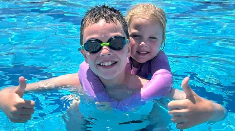 Handout A boy in goggles smiles and does a thumbs up at the camera, he is in blue water, a girl in a pink top smiles, arms around his neck.