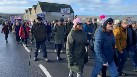 A line of people walking on the coast, a few holding banners 