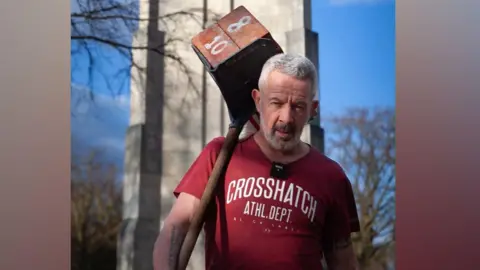 A man with grey hair in a red t-shirt, holding a wooden pole and a carrier full of bricks over his shoulder, in front of a white memorial tower. Around is blue sky and trees.