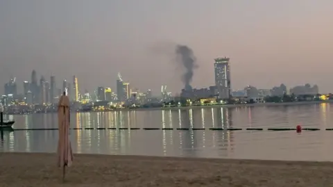 Reuters/Handout A city skyline featuring large skyscraper buildings. The photograph has been taken from a beach with an area of sand and water visible. Smoke can be seen rising from an area of buildings following a missile strike.
