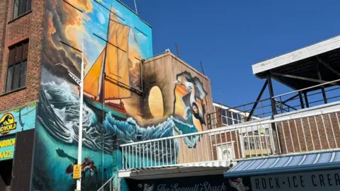 A large-scale, colourful wall painting showing a traditional sailing boat with large orange sails battling through large waves in a blue sea. The sky depicts a storm clearing. A lobster, seabirds and a puffin can also be seen. It is painted on the side of a large brick building under a blue sky. In the bottom right of the photo, shops can be seen with signs reading "The Seagull Shack", and "Rock, Ice Cream".