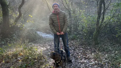 Christian Jones wearing a puffer coat and a bobble hat stands in a wooded clearing next to a stream. He is holding the lead of his Staffordshire terrier Bud. There are visible sunbeams coming from over Christian's right shoulder. 