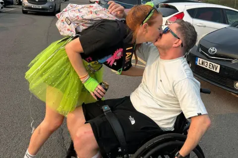 Anna Thomas A man with brown hair wearing a white t shirt, black shorts and sunglasses, sat in a wheelchair in a car park with cars in the background. A woman with red hair, dressed in a black t shirt and neon green tutu is leaning down and kissing him.