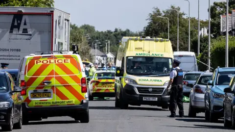 A police van, an ambulance and a police car on Hart Street in Southport on the day of the knife attacks on 29 July 2024. An area is cordoned off with police tape and a policeman is standing in the road next to a row of parked cars.