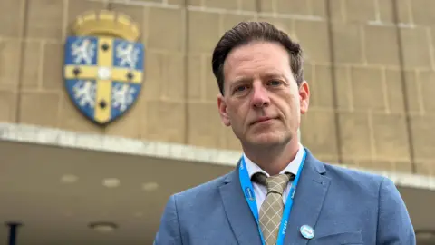 New Durham County Council leader Andrew Husband outside County Hall. He is wearing a light blue suit jacket, a white shirt and a gold tie. His brown hair is swept back.