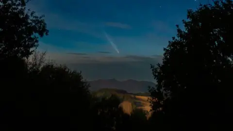 Tomahawk/BBC Weather Watchers Comet A3 appearing above hills and fields and framed by trees.