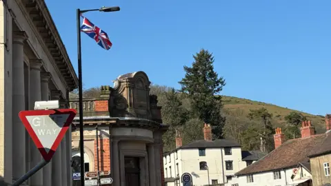 Charlie Stubbs / BBC Here shows a skyline of Church Stretton - with a big green hill. It also shows buildings in the foreground, with old stonework and white and red brick buildings. There is also a flag of the United Kingdom tied up to the top of a black lampost, and a red 'Give Way' traffic sign.