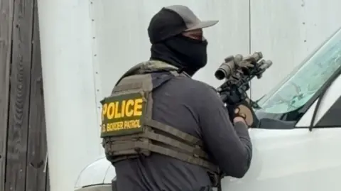 Zoë Higgins Z tyłu widać lekko odwróconego mężczyznę w czarnej masce, okularach i kamizelce kuloodpornej, trzymającego broń z lunetą. Pochyla się nad dużym białym samochodem.