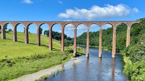 Mark Grierson A viaduct with tall red-brick arches runs down a grassy hill and over a river and into a green forest on a bright and sunny day.