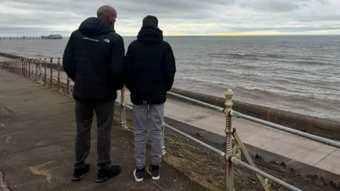 A man wearing a black North face jacket stands next to a teenager with grey trousers looking out to sea on a grey day. 