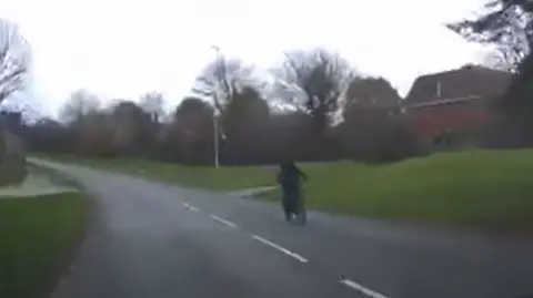 Northamptonshire Police A slightly blurred image of a road, with a man on a bike, with a red brick house to the right and grass verges on either side of the road. There are trees on both sides of the road. 
