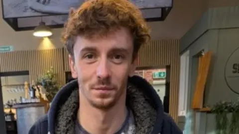 North Yorkshire Police The image shows a man with light brown untidy hair and facial stubble on a thin white face. He is seated at a table in a café. He is wearing a dark zip‑up hoodie over a patterned T‑shirt.