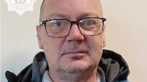 A mugshot of a bald man with rectangular glasses. He is staring into the camera and has stubble around his chin and upper lip. The hood of a dark grey jumper can be seen at the bottom of the image.