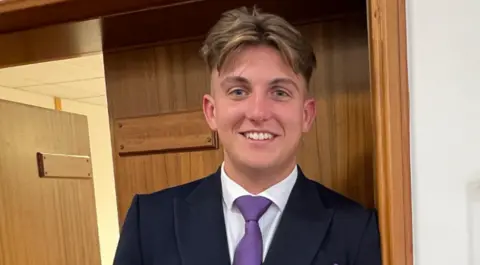 A man in a dark jacket, purple tie and white shirt is next to a door frame.