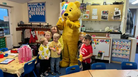Pudsey bear pictured with a group of small children in a classroom. He has both thumbs up. The children are smiling and some of them are looking at him.
