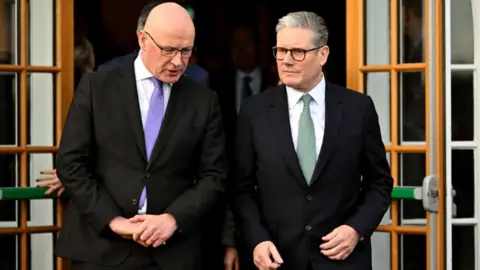 John Swinney, who is bald with glasses and Sir Keir Starmer, grey-haired with glasses, walk through open pane glass doors. Swinney is on the left and Starmer is on the right. Swinney, with his head slightly bowed, is speaking. Both are wearing black suits and white shirts. Swinney is wearing a purple tie, Sir Keir a green tie. 