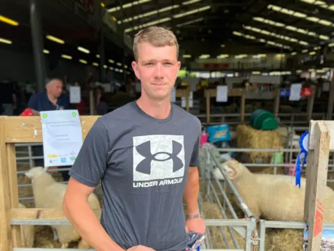 BBC Young man in a t-shirt standing in front of a showground barn with sheep pens