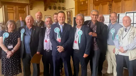 Reform candidates, 10 men and one woman, pose together for a photo in inside a building, following the local election results. They are all wearing a party rosette and looking at the camera smiling. 