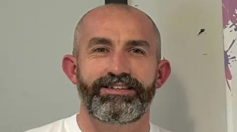 Gerard Donaghy is standing in front of a white wall, wearing a white t-shirt and is smiling at the camera. He has a bald head and a dark beard with grey hair through it.