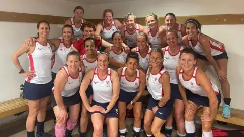 The England women's over-45 squad for the Hockey Masters World Cup. A group of women are all huddled together, many with their arms round each other, and looking at the camera and smiling. Some of them are stood on a wooden bench. They are all wearing hockey kit, which consists of a white vest with red details and navy blue shorts. 