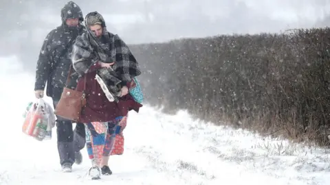 A cold-looking man and woman walk along a snow-covered path beside a hedge in a blizzard. They are wearing winter coats and carrying groceries including toilet roll.