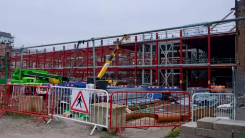 A construction site behind red and white metal fences. Two vehicles are parked next to a pile of girders and other building materials. The red and silver shell of a building can be seen in the background,