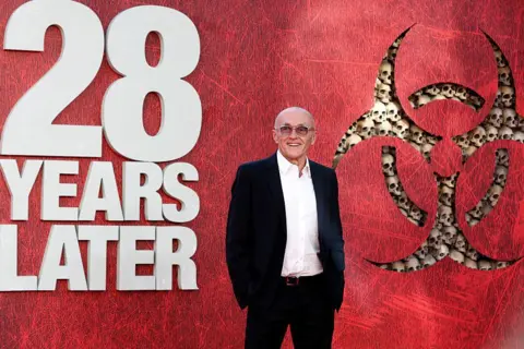 Getty Images Director Danny Boyle smiling for pictures against an advertising board for 28 Years Later, wearing a dark suit jacket, white shirt and black trousers