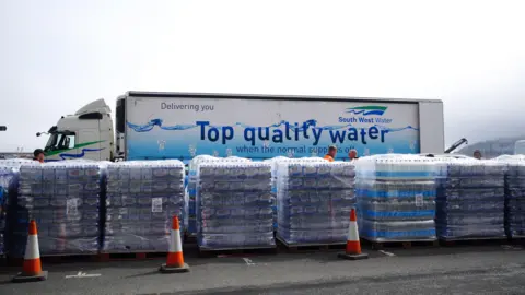 Six big packages of bottled water in a car park. Behind them is a lorry with South West Water branding and the line: Delivering you top quality water.