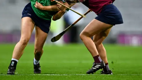 Getty Images Two players in navy skorts and black trainers playing camogie on a grass pitch.