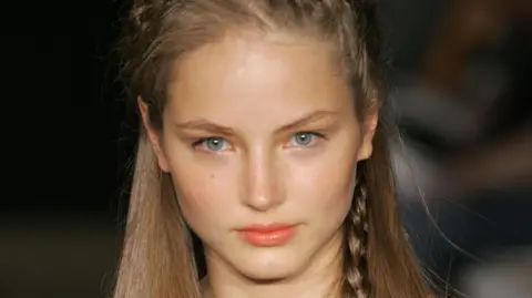 Getty Images Ruslana Korshunova, a young woman with fair hair, part of which is braided and tucked behind her ear, looks straight at the camera in a close-up shot taken during a catwalk show. Her face is starkly lit with the background in darkness.