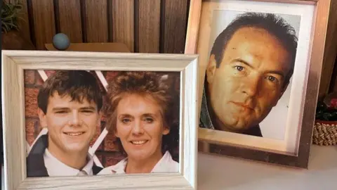 Duas molduras para fotos estão posicionadas no topo de uma lareira, uma contendo uma fotografia de Sheila e Barry Grant sorridentes. O outro é de Jimmy Corkhill.