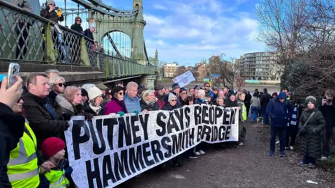 Dozens of people stand beside Hammersmith Bridge holding a large banner reading “Putney Says Reopen Hammersmith Bridge,” with others watching from the bridge and nearby path.