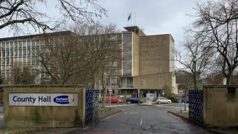 Tall brick building with sign at on the gate at the front saying "Durham County Council: County Hall".