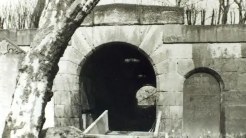 A black and white stone archway that opens up a dark tunnel. There is a grave to the right of it and numerous trees in front of it and behind it.