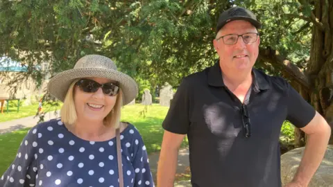 Glenda Grey is smiling and has blonde hair. She is wearing a hat, sunglasses and a blue and white polka-dot top. On the right, Phil Grey is wearing glasses, a black polo shirt and baseball cap. Behind them there is a graveyard. It is sunny.