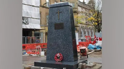 Cumberland Council The war memorial in its new location in Market Square. The monument is a black obelisk, with a cross placed near its summit. At its base is a red poppy wreath. Behind you can see the construction works.