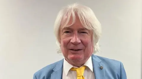 Arun District Council A man with collar length grey hair is looking at the camera. He is wearing a pale blue suit, white shirt and yellow tie