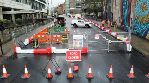 A road that has been sealed off with fencing and traffic cones, as well as a Road Closed sign. Workers in orange high vis jackets can be seen in the background.