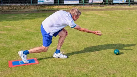 Craig McKernon A man in a white shirt and blue shorts bowling on a green.