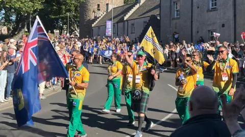 BBC Athletes walk down the middle of the road waving flags in tracksuits. Crowds cheer at the side of the road.