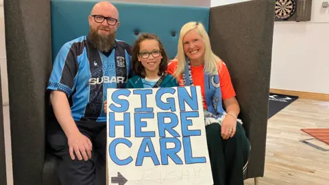 Leo is holding his sign which reads 'Sign Here Carl and features the player's signature at the bottom. Leo is flanked by his dad Paul, who is wearing a retro Coventry City top and his mum Clare, who has the club's hot coral away kit on. She also has a Coventry City scarf around her neck.