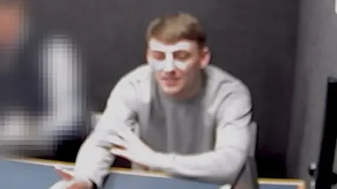 Kane Farragher is in a grey police interview room and is speaking with someone off-camera with one arm outstretched and the other crossed over his body. He has short brown hair and is wearing a grey jumper. There is someone in blue sitting beside him, but they are blurred out.